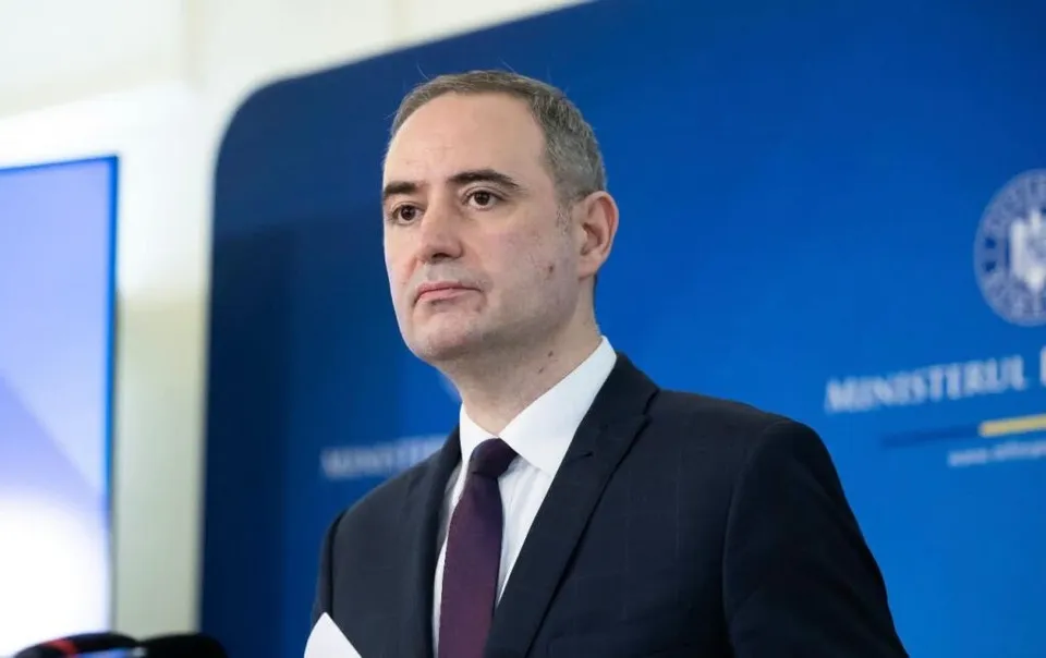 Un bărbat cu părul scurt, purtând sacou închis la culoare, cămașă albă și cravată purpurie, stă în fața unui fundal albastru cu inscripții albe.