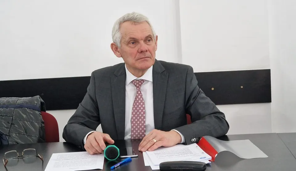 Un bărbat cu părul alb, purtând sacou gri și cravată roz, stă la o masă cu hârtii și ochelari în față.