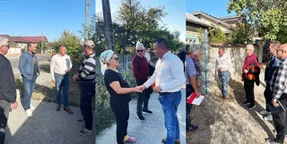 Un grup de persoane stă pe un drum asfaltat lângă o clădire, în timp ce un bărbat în cămașă albă discută cu o femeie.