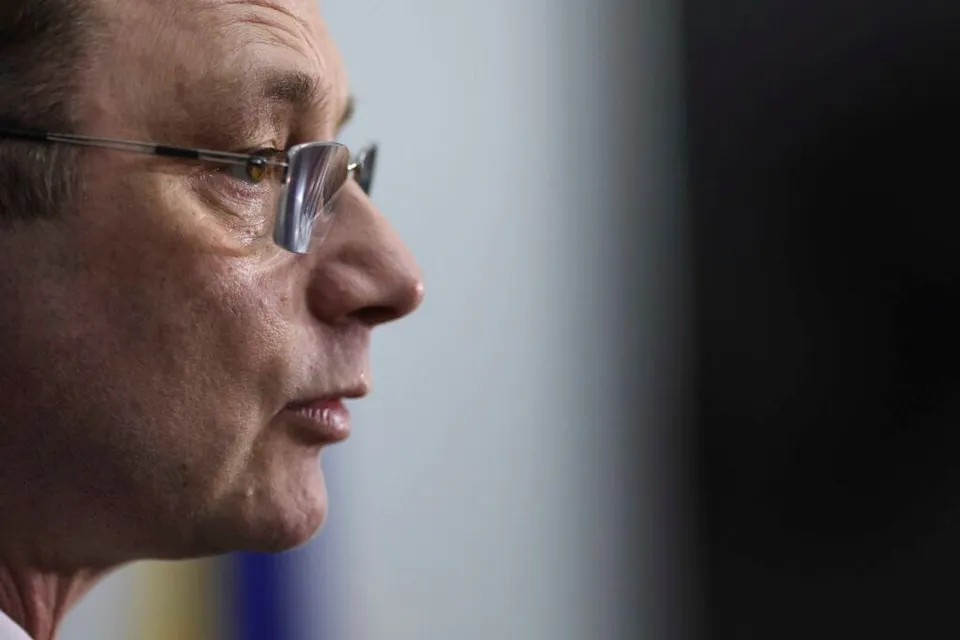 Un bărbat cu ochelari este surprins în profil, având fața orientată spre dreapta pe un fundal neclar și deschis la culoare.