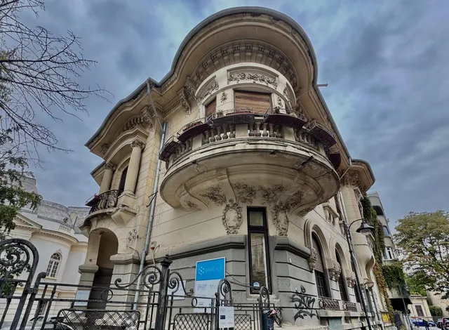 O clădire cu arhitectură eclectică prezintă un balcon circular susținut de elemente decorative și ferestre înalte sub un cer înnorat.