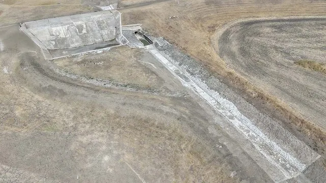 O vedere aeriană prezintă o structură de beton gri situată într-un peisaj arid cu terenuri cultivate și poteci de pământ.