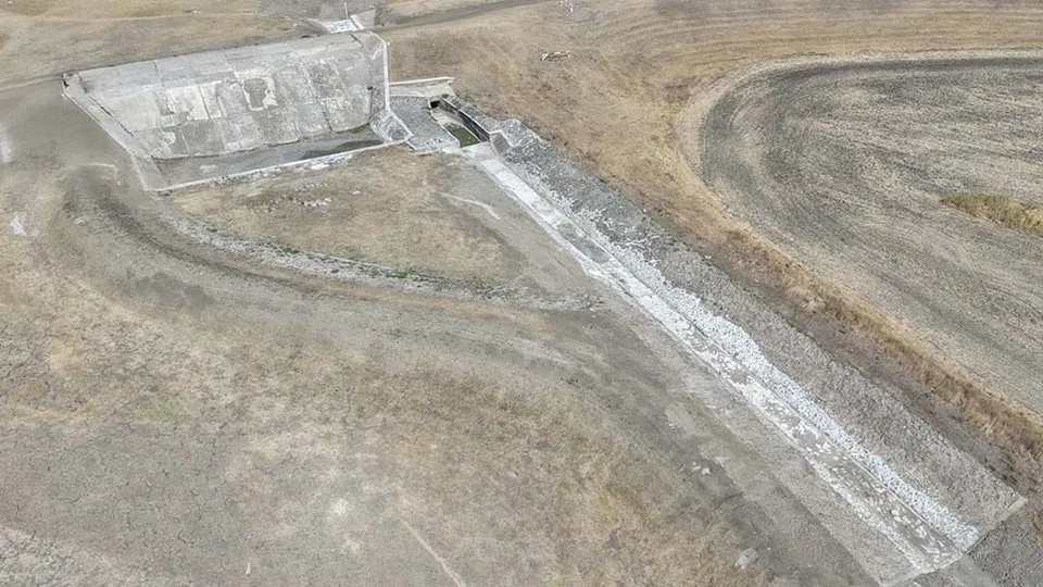 O vedere aeriană prezintă o structură de beton gri situată într-un peisaj arid cu terenuri cultivate și poteci de pământ.