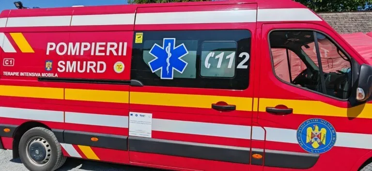 Un vehicul de serviciu roșu cu inscripții de pompieri și SMURD este parcat pe o suprafață asfaltată sub cerul senin.