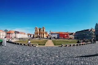 O piață pavată cu pietriș prezintă bănci albe, un gard jos și o zonă cu iarbă în fața unor clădiri colorate sub un cer senin.