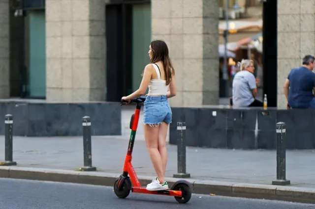 O femeie cu părul lung, purtând un top alb și pantaloni scurți din denim, stă pe o trotinetă electrică roșie pe marginea unui trotuar.