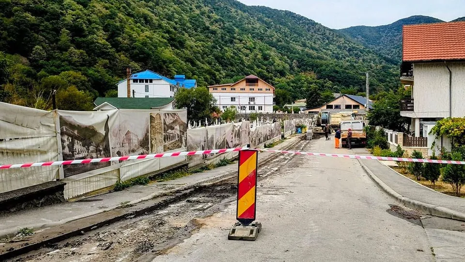 O stradă asfaltată este delimitată de un gard de protecție și bandă de semnalizare, având un con de trafic galben și roșu în prim-plan.