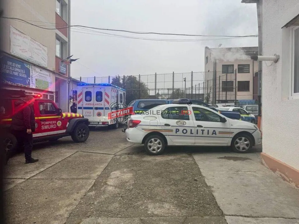 Un vehicul de poliție alb și un vehicul de ambulanță sunt parcate pe un teren de beton lângă clădiri, în prezența unei mașini de pompieri roșie.