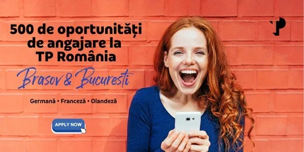 O femeie cu părul roșcat, zâmbind cu gura deschisă, ține un telefon mobil în fața unui zid de cărămidă roșie pe care scrie un text publicitar.