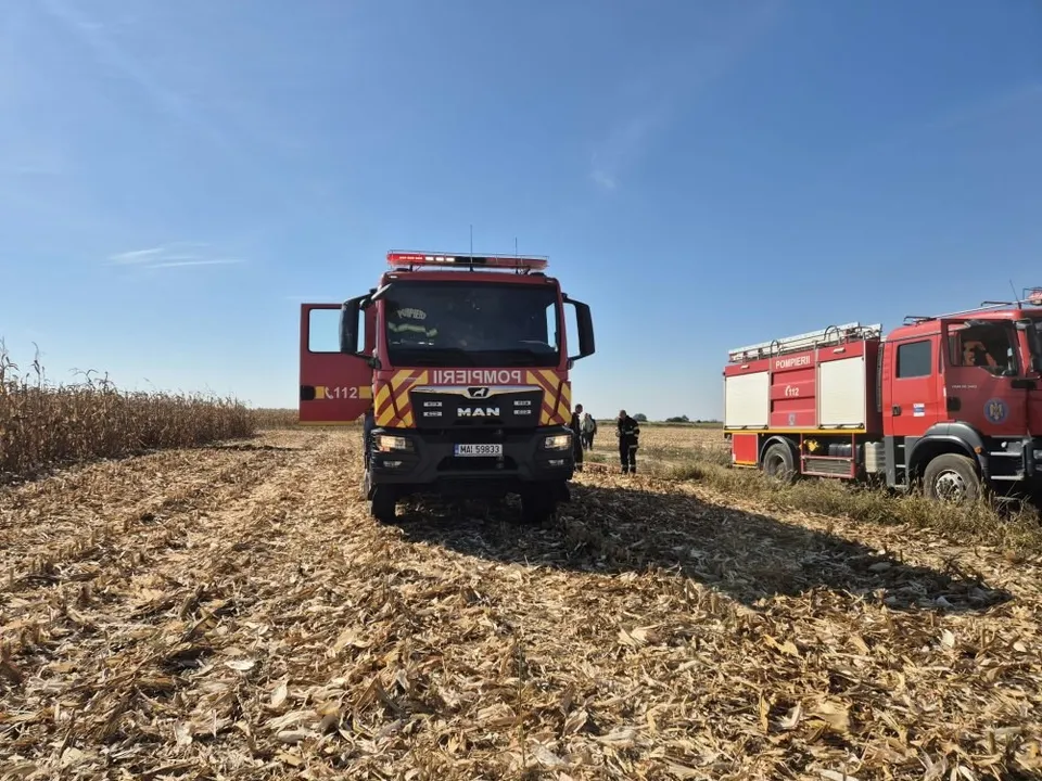 Două autospeciale de pompieri roșii sunt parcate într-un câmp cu resturi de plante uscate sub un cer senin.
