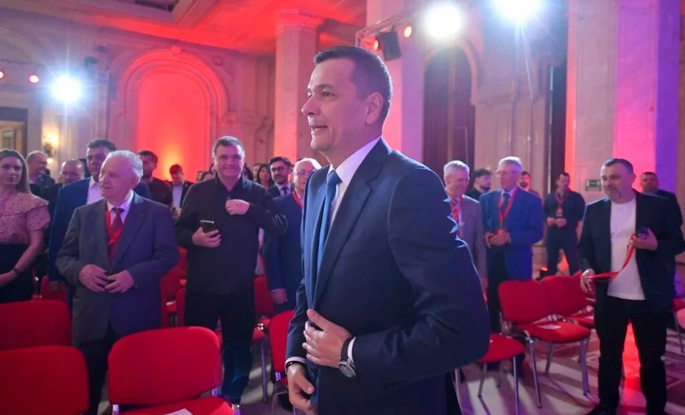 Un bărbat într-un costum închis și cămașă albă stă în prim-plan, în timp ce alți bărbați în costume stau în spatele lui într-o încăpere iluminată în roșu.