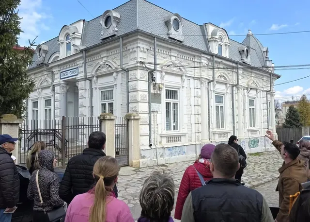 Un grup de persoane stă cu spatele la cameră în fața unei clădiri albe cu detalii arhitecturale și ferestre înalte sub un cer înnorat.