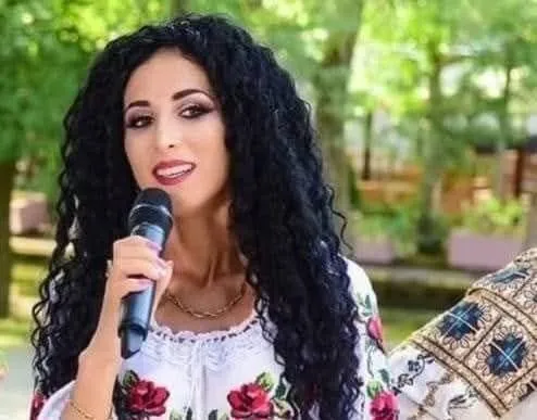 O femeie cu părul negru și creț ține un microfon negru în fața feței, purtând o bluză albă cu motive florale roșii.