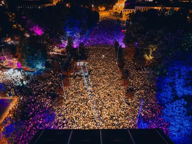 O mulțime de oameni este adunată într-o zonă deschisă, iluminată de lumini colorate de nuanțe de albastru și roz, sub un cer de noapte.