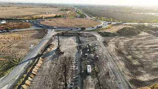 O vedere aeriană prezintă un șantier cu utilaje grele, un drum de pietriș și o intersecție circulară înconjurată de terenuri deschise.