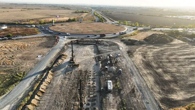O vedere aeriană prezintă un șantier cu utilaje grele, un drum de pietriș și o intersecție circulară înconjurată de terenuri deschise.