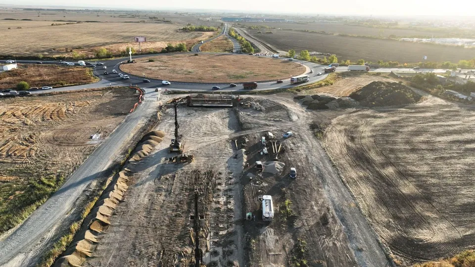 O vedere aeriană prezintă un șantier cu utilaje grele, un drum de pietriș și o intersecție circulară înconjurată de terenuri deschise.