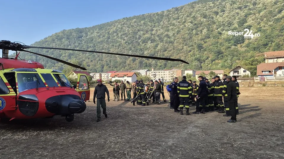 Un elicopter roșu și galben stă pe un teren deschis lângă un grup de persoane purtând uniforme de serviciu și un deal împădurit.