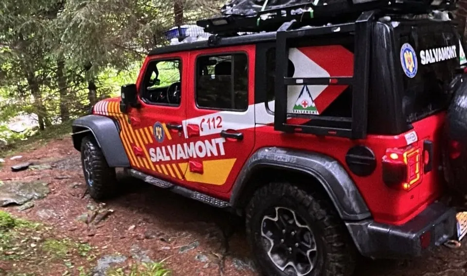 Un vehicul de salvamont roșu și alb cu inscripția SALVAMONT este parcat într-o zonă împădurită lângă un drum de pământ.