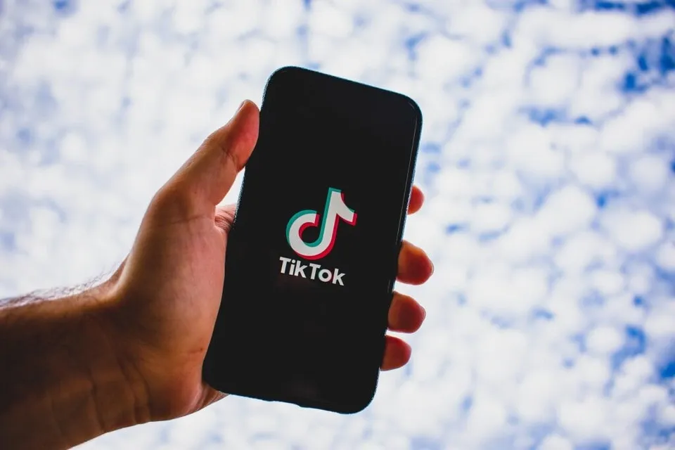 O mână ține un smartphone negru care afișează logoul TikTok pe un fundal de cer cu nori albi.