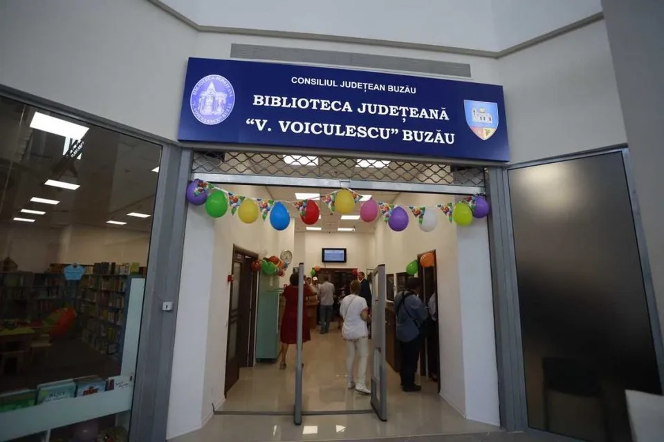 O intrare de clădire cu un panou albastru deasupra, decorată cu baloane colorate, unde câțiva oameni intră într-un spațiu iluminat.