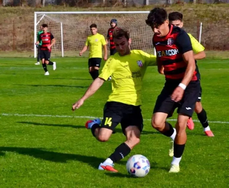 Un jucător în echipament galben lovește un balon pe un teren de fotbal verde, fiind urmărit de un adversar în echipament roșu și negru.