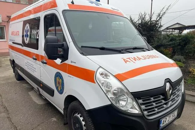 Un vehicul de tip ambulanță alb cu dungi portocalii este parcat pe un drum asfaltat lângă o clădire și vegetație.