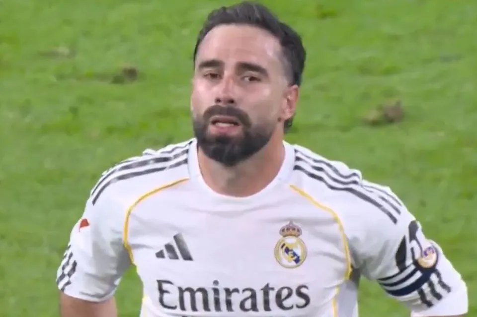 Un bărbat cu barbă poartă un tricou alb cu dungi negre pe umeri și logoul Emirates pe piept, stând pe un teren de iarbă verde.