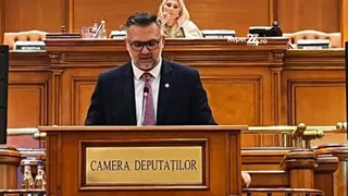 Un bărbat cu ochelari și cravată roz stă în spatele unui podium de lemn pe care scrie Camera Deputaților într-o încăpere cu mobilier din lemn.