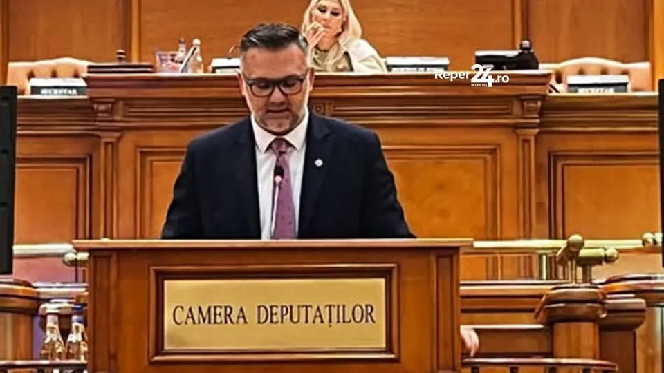 Un bărbat cu ochelari și cravată roz stă în spatele unui podium de lemn pe care scrie Camera Deputaților într-o încăpere cu mobilier din lemn.