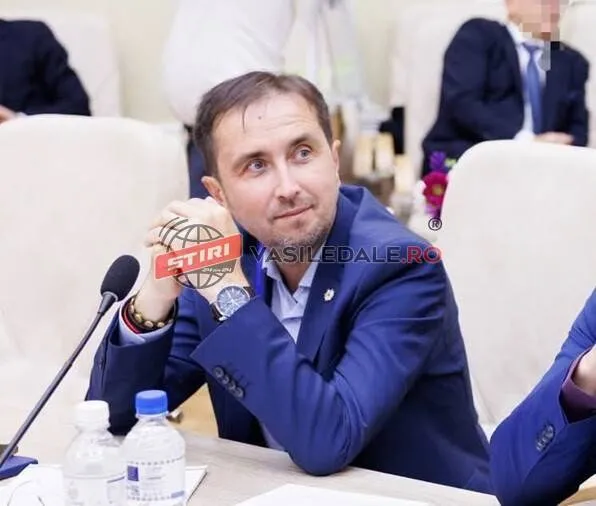 Un bărbat cu sacou albastru stă la o masă cu microfoane și sticle de apă în fața unor scaune albe.