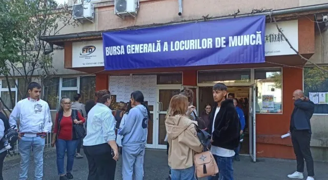 Un grup de oameni stă în fața unei clădiri sub un panou albastru pe care scrie text alb.