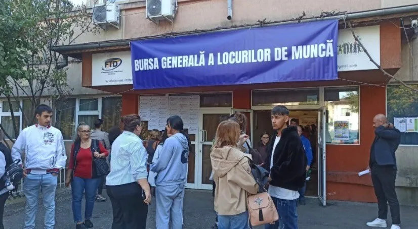 Un grup de oameni stă în fața unei clădiri sub un panou albastru pe care scrie text alb.