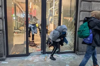 O persoană cu hanorac gri este aplecată lângă o vitrină de sticlă spartă, în timp ce un alt individ merge pe trotuar purtând un rucsac verde.
