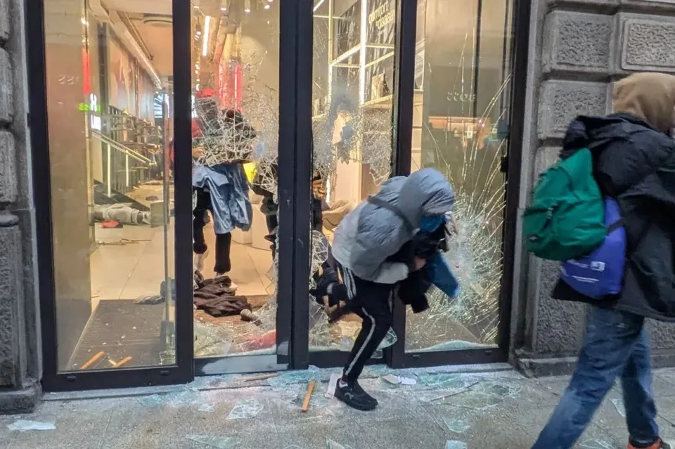 O persoană cu hanorac gri este aplecată lângă o vitrină de sticlă spartă, în timp ce un alt individ merge pe trotuar purtând un rucsac verde.
