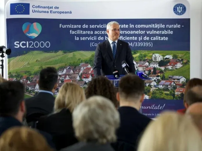 Un bărbat într-un costum închis la culoare stă în spatele unui microfon în fața unui panou informativ cu un peisaj rural.