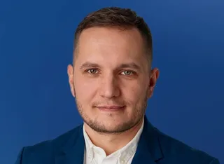Un bărbat cu părul scurt și barbă scurtă poartă un sacou albastru peste o cămașă albă, fiind fotografiat în fața unui fundal albastru.