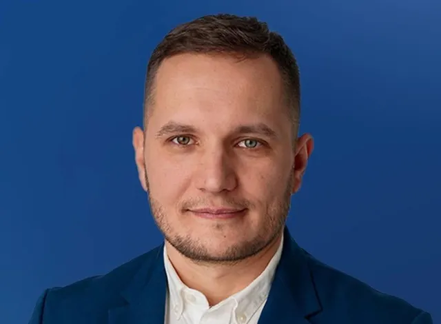 Un bărbat cu părul scurt și barbă scurtă poartă un sacou albastru peste o cămașă albă, fiind fotografiat în fața unui fundal albastru.