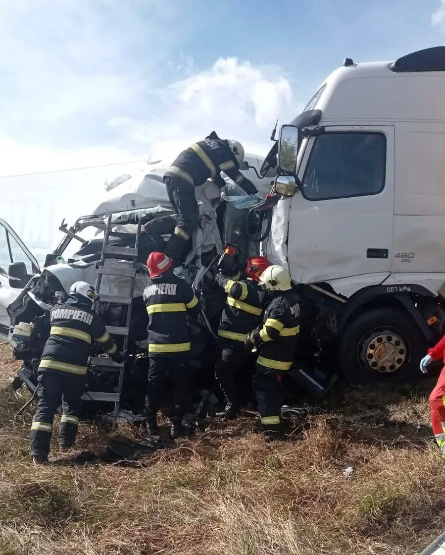 Mai mulți pompieri în uniforme de lucru în negru și galben intervin lângă un microbuz alb și un camion alb pe un teren cu iarbă.