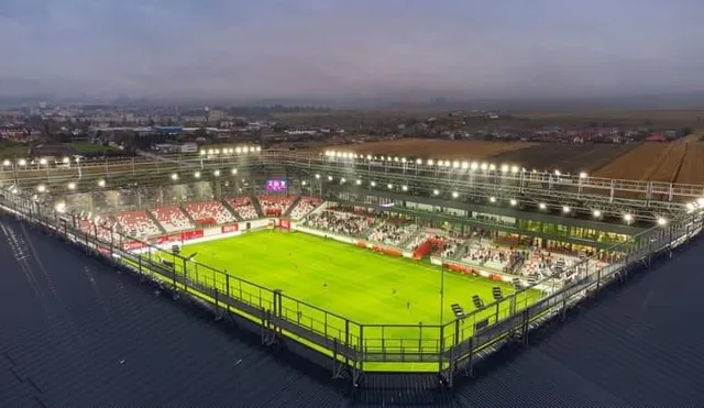 O vedere aeriană a unui stadion de fotbal iluminat cu un teren verde înconjurat de tribune cu scaune roșii și albe sub un cer închis.
