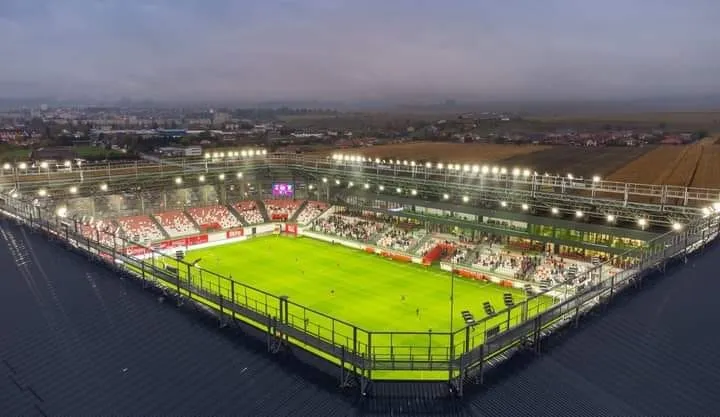 O vedere aeriană a unui stadion de fotbal iluminat cu un teren verde înconjurat de tribune cu scaune roșii și albe sub un cer închis.