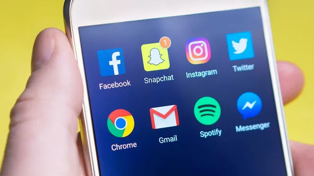O mână ține un smartphone care afișează diverse pictograme de aplicații pe un fundal albastru, inclusiv Facebook, Snapchat, Instagram și Twitter.