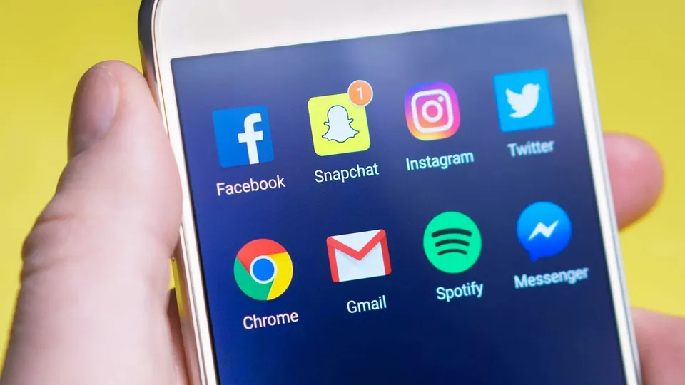 O mână ține un smartphone care afișează diverse pictograme de aplicații pe un fundal albastru, inclusiv Facebook, Snapchat, Instagram și Twitter.