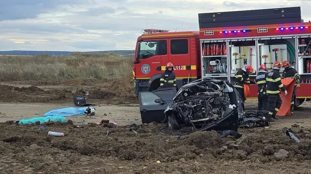 Un autoturism negru distrus stă pe un teren de pământ lângă un camion roșu și mai mulți pompieri în uniforme de lucru.