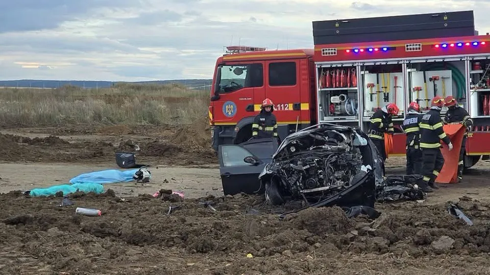 Un autoturism negru distrus stă pe un teren de pământ lângă un camion roșu și mai mulți pompieri în uniforme de lucru.