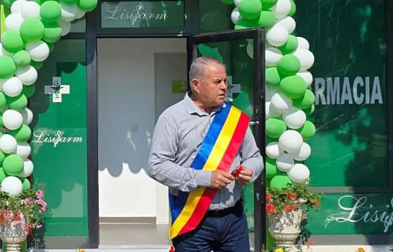 Un bărbat în vârstă, cu o eșarfă tricoloră pe umăr, stă într-o intrare decorată cu baloane verzi și albe, în fața unei farmacii.