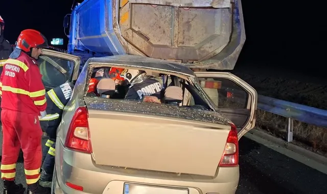 Un pompier în echipament roșu și galben stă lângă un autoturism argintiu cu portierele deschise, lângă o autobasculă răsturnată pe partea laterală noaptea.