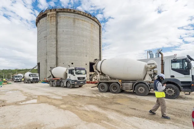 Două camioane cu betoniere albe sunt parcate pe un teren de construcții lângă un cilindru de beton înalt sub un cer înnorat.