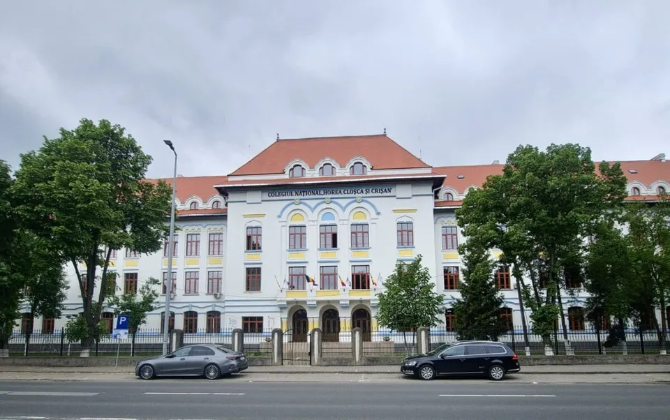 O clădire albă cu acoperiș roșu, ferestre multiple și un inscripție pe fațadă, situată în spatele unor copaci verzi și a unui drum asfaltat.