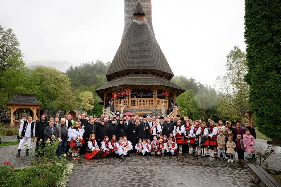 Un grup mare de oameni, unii purtând costume populare, stau grupați în fața unei clădiri din lemn cu acoperiș înalt și conic.